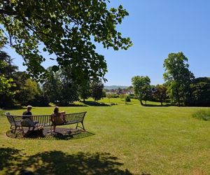 Two people sitting on a wide curved bench overlooking the vast lawns of Hatherley Park with trees on either side