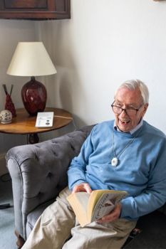 Elderly man sitting on his sofa at home, reading a book, wearing a lifeline pendant