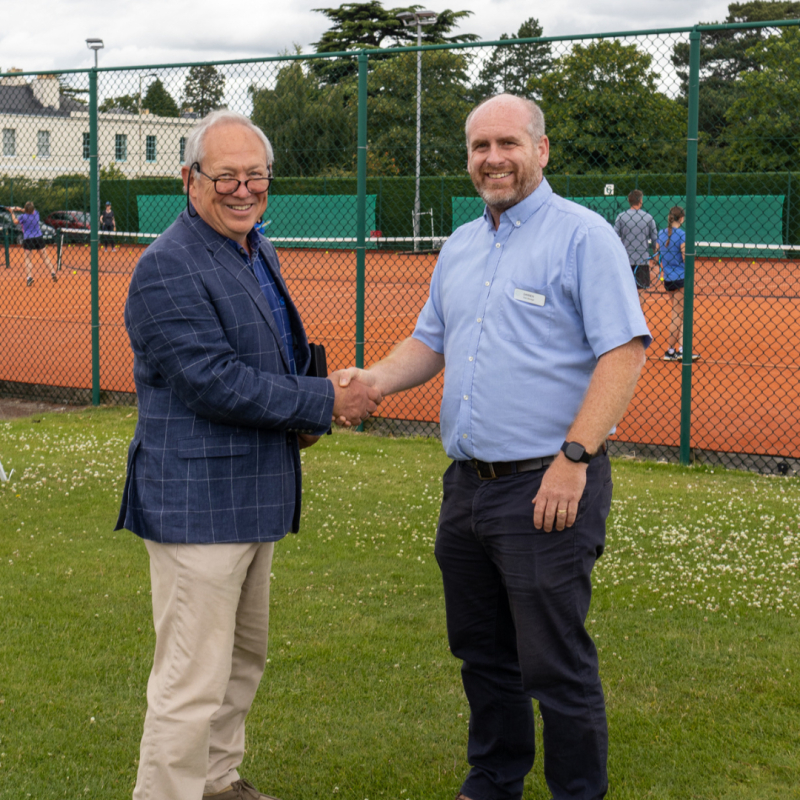 Cllr Iain Dobie shakes hands with Darren Morris from the East Glos club. They are stood in front of the tennis courts at the club.